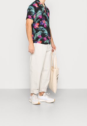 Korte mouwen zwarte shirt met een levendige tropische bloemenprint. Lichtgekleurde losse broek en witte sneakers. Beige tote bag in de hand.