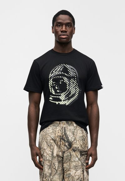 Μαύρο βαμβακερό T-shirt με μεγάλο γραφικό ενός στυλιζαρισμένου προσώπου με ρίγες και σχέδια. Συνδυασμένο με μπεζ παντελόνι με σχέδια.