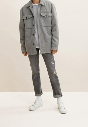 Chaqueta de camisa de mezcla de lana gris con dos bolsillos en el pecho, llevada sobre una camiseta de manga larga blanca con textura, combinada con jeans gris desgastados y zapatillas blancas.
