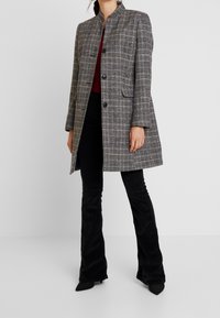 Grijze geruite jas met een mandarin-kraag, sluiting met één knoop en klepzakken, gecombineerd met zwarte flared broek en puntige hakken.