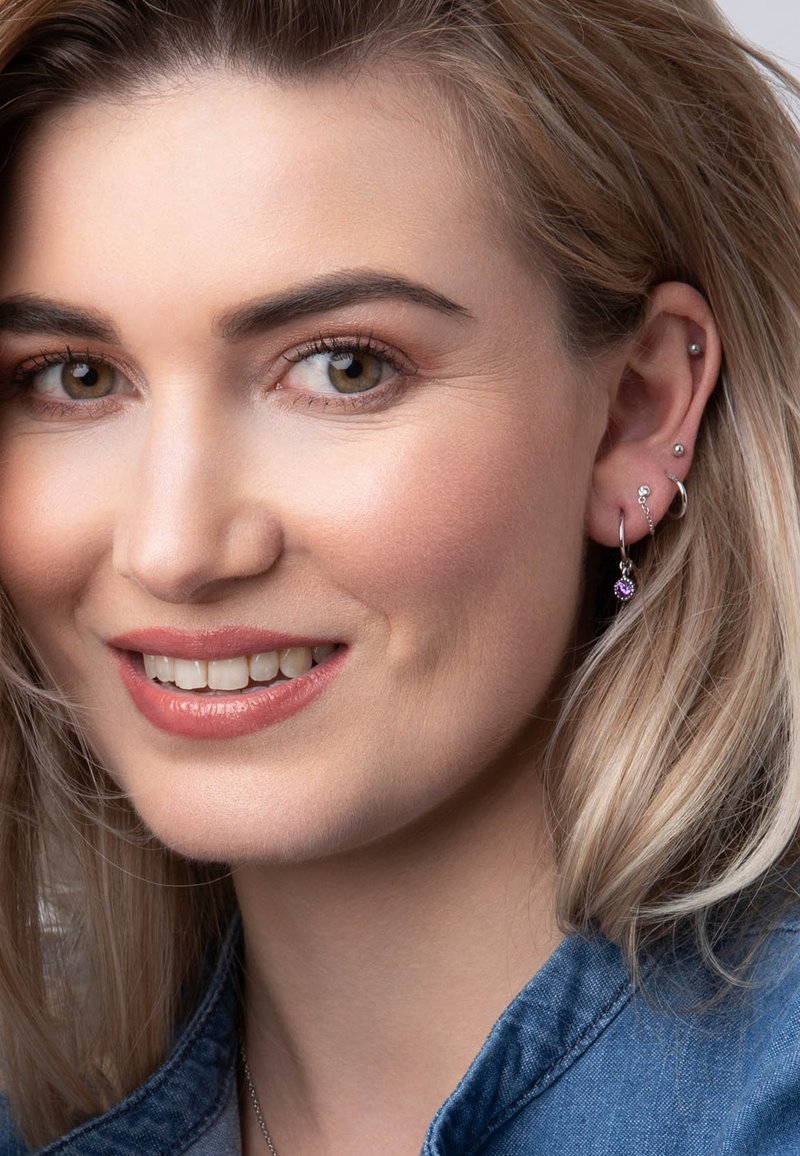 Zilveren haak oorbellen met paarse edelsteenaccenten, gecombineerd met verschillende studs. Het model heeft licht haar en draagt een blauwe denim top.