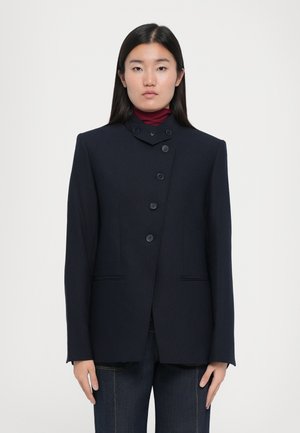 Veste tailleur bleu marine avec col haut, fermeture frontale à cinq boutons et deux poches avant. Texture de tissu lisse et silhouette structurée.