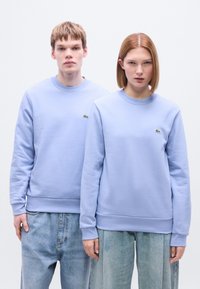 Ανοιχτό μοβ φούτερ με ριμπ μανσέτες και τελείωμα, με μικρό πράσινο λογότυπο Lacoste. Φορεμένο με ανοιχτό μπλε τζιν παντελόνι.