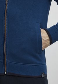 Marine blå zip-hettegenser laget av myk stoff, med en gullglidelås, sidelommer, og ribbestrikkede mansjetter og kant med en kontrasterende kantdetalj.