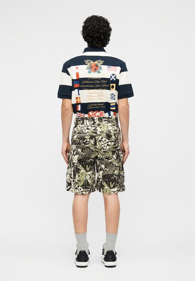 Άνδρας στέκεται με την πλάτη, φορώντας μπλε-σκούρο και λευκό κοντομάνικο πουκάμισο με διεθνείς σημαίες και φορμά καμουφλάζ.