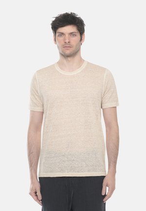 Uomo con capelli corti e scuri che indossa una maglietta beige tinta unita a maniche corte e pantaloni neri, in piedi di fronte a uno sfondo bianco.