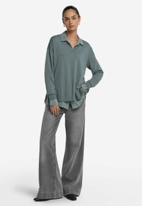 Kvinna i en lös grågrön tröja över en kragskjorta, med vida grå jeans och svarta skor, stående med en hand i fickan.