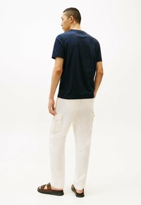 T-shirt a maniche corte color navy, abbinato a pantaloni cargo leggeri beige con tasche. Il modello è in piedi su uno sfondo neutro, indossando sandali marroni.