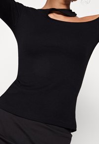 Frau trägt ein enganliegendes, schwarzes Rippentop mit einem asymmetrischen Ausschnitt an der rechten Schulter vor einem weißen Hintergrund.