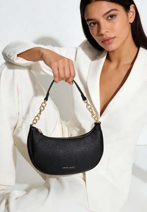Donna in abito bianco che tiene una borsa nera testurizzata con tracolla a catena dorata e logo "pierre cardin", posando su sfondo bianco.