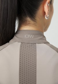 Grå treningsskjorte med et perforert panel på baksiden og en krage med "ICIW" subtilt trykt nedenfor. Glatt tekstur og strømlinjeformet design.