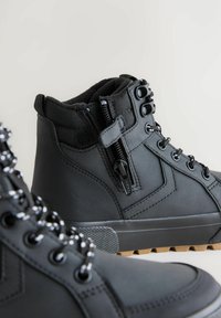 Schwarze hochhackige Stiefel mit Schnürsenkeln und einem seitlichen Reißverschluss, aus strukturiertem Stoff und mit einer Gummisohle. Der Hintergrund ist einfarbig und hell.