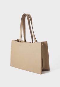 Sac fourre-tout en cuir beige avec deux longues poignées, de forme rectangulaire et au design minimaliste, mettant en avant des coutures discrètes et des accents dorés.
