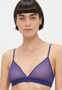 Vrouw met kort donker haar die een doorschijnende paarse bralette met dunne bandjes draagt, kijkt recht vooruit tegen een effen achtergrond.