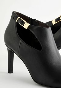 Stivaletti neri in pelle fino alla caviglia con un design a taglio, caratterizzati da un accentuato bottone dorato e un alto tacco a stiletto. Texture liscia e forma elegante.
