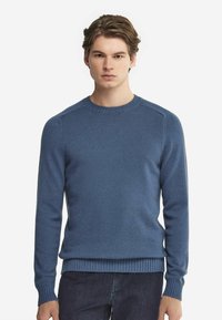 Blå stickad tröja med rund halsringning, ribbade ärmslut och fåll. Tyget verkar mjukt med en slät textur, figursydd design.