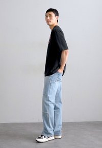 Zwarte graphic t-shirt, losse lichtblauwe jeans en zwart-witte sneakers. Model staat zijwaarts tegen een grijze achtergrond.