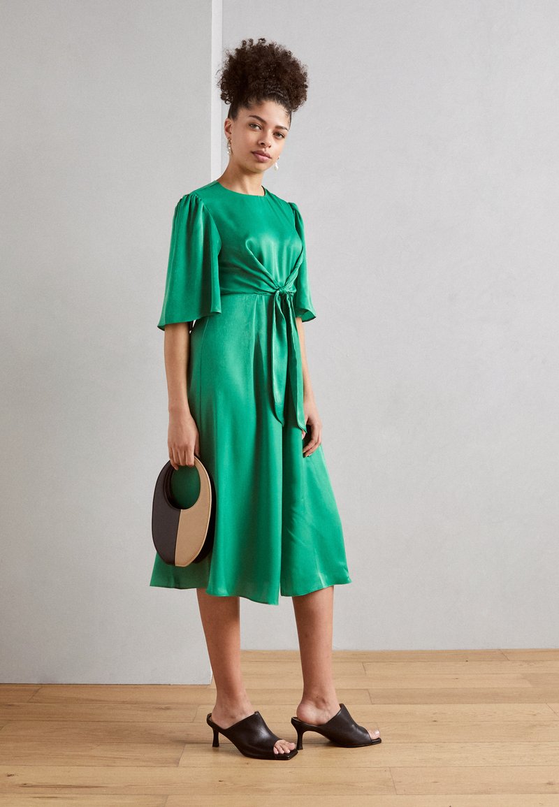 Grünes Satin-Kleid mit gebundenem Taillengürtel, kurzen Puffärmeln und ausgestellt geschnittenem Saum. Kombiniert mit schwarzen offenen Mules und einer zweifarbigen runden Handtasche.