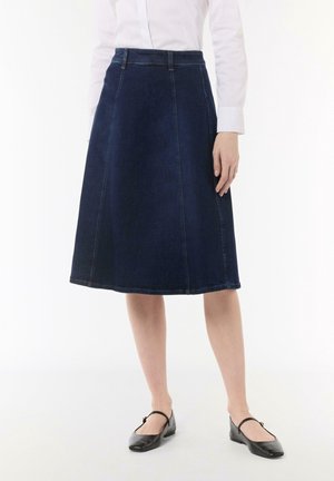 Mujer con una falda vaquera azul oscuro hasta la rodilla, camisa blanca de manga larga y zapatos planos negros con correas, de pie sobre un fondo blanco.