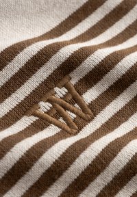 Ύφασμα με ριγέ καφέ και εκρού χρώμα και απαλή υφή· διαθέτει κεντημένο λογότυπο σε καφέ κλωστή, τοποθετημένο στη κάτω αριστερή περιοχή.