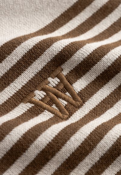 Tissu rayé marron et crème avec une texture douce ; présente un logo brodé en fil marron, positionné dans la partie inférieure gauche.