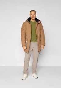 Brun pufferjakke med svart hette, grønn t-skjorte, lysebeige bukser og nøytrale sneakers. Jakken har glidelås og trykknapplukking.