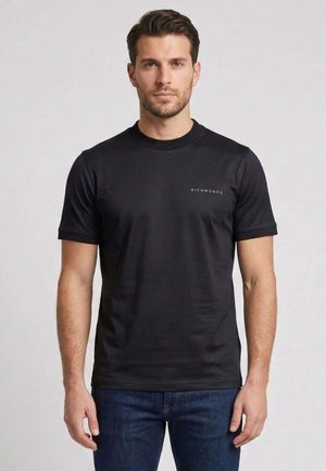 Uomo con capelli castani corti che indossa una maglietta nera semplice RichmondX e jeans blu scuro, in piedi davanti a uno sfondo neutro.