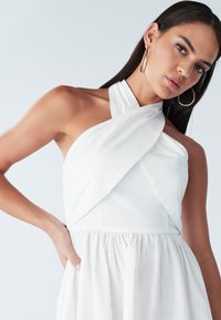 Biele šaty s halterneckom, zdobené obtiahnutým živôtikom, nariaseným pásom a jemnou, ľahkou látkou. Ruka modelky spočíva v bočnom vrecku.