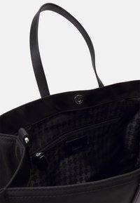 Schwarze Tragetasche mit glatter Oberfläche, doppelten Schultergurten und einem Innenleben mit Hahnentrittmuster sowie einer Reißverschlusstasche. Magnetverschluss.