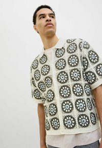 Pletena majica s kratkimi rokavi, ki ima kremasto osnovo z zapletenimi krožnimi čipkastimi vzorci v temno zeleni in modri barvi, dopolnjena z gumbom za zapiranje.