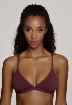 Frau mit lockigem Haar trägt ein dreieckiges, dunkelrotes Bustier und blickt geradeaus vor einem schlichten hellgrauen Hintergrund.