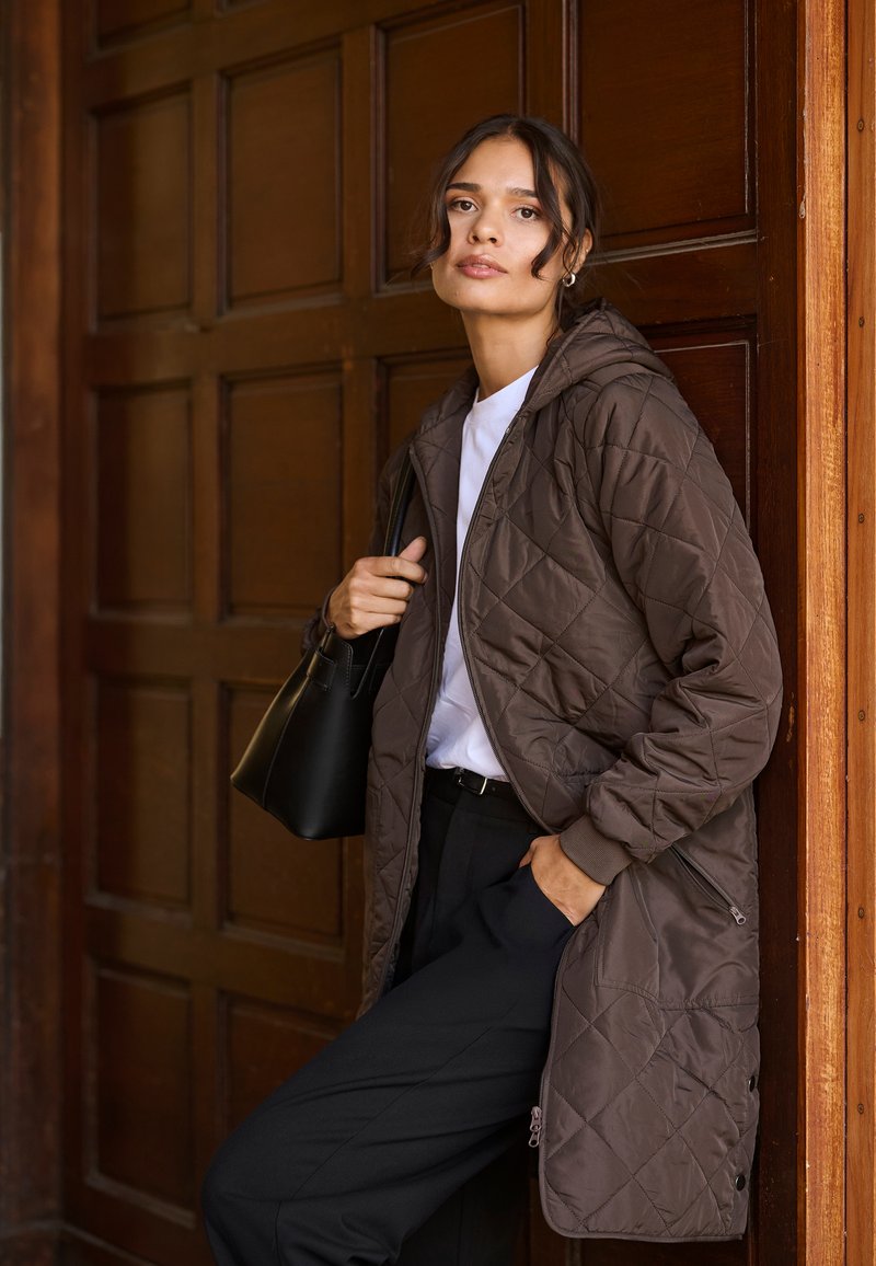 Abrigo acolchado marrón con capucha, con bolsillos con cremallera y una textura suave, combinado con una camisa blanca y pantalones negros.