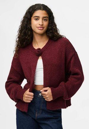 Gilet en tricot bordeaux avec un col rond, des poignets épais à côtes et une fermeture à bouton floral. Porté sur un haut blanc et un pantalon en denim.