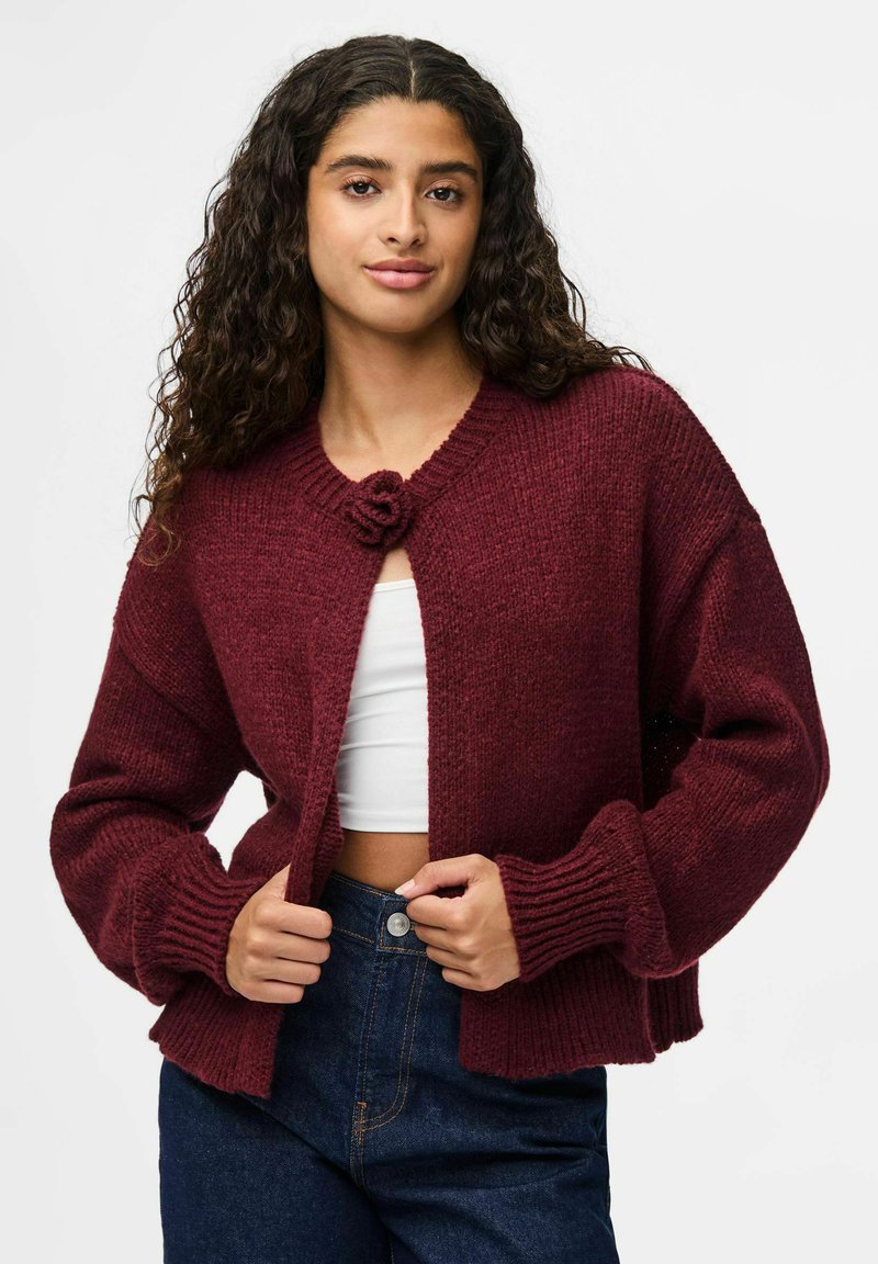 Bordeaux gebreide cardigan met een ronde hals, dikke geribbelde manchetten en een bloemenknopen sluiting. Draagt over een witte top en denim broek.
