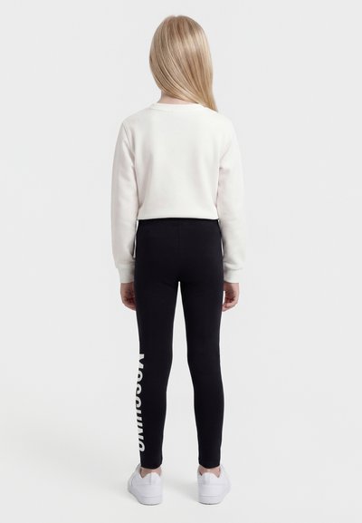 Laps, kellel on pikad blondid juuksed, seisab seljaga vaataja poole, kandes valget dressipluusi, musti retuuse, mille vasakul säärel on vertikaalne valge tekst, ning valgeid tossusid.