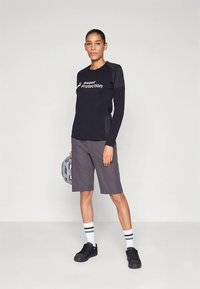 Μαύρο μακρυμάνικο πουκάμισο με το λογότυπο «Sweet Protection», γκρι σορτς, μαύρα αθλητικά παπούτσια και ριγωτές λευκές κάλτσες. Κρατά ένα γκρι κράνος.