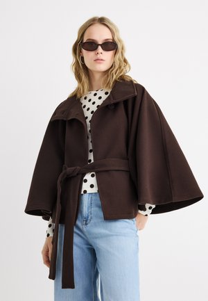 Γυναίκα που φοράει σκούρο καφέ παλτό-μανδύα με ζώνη πάνω από λευκή μπλούζα με πουά και ανοιχτόχρωμα μπλε τζιν, με σκούρα γυαλιά ηλίου και κρίκους αυτιών.