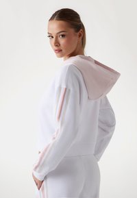 Witte cropped sweatshirt met een capuchon in een roze accent. Kenmerkt zich door drie verticale strepen in roze en beige op de mouwen. Zachte textuur, relaxed fit.