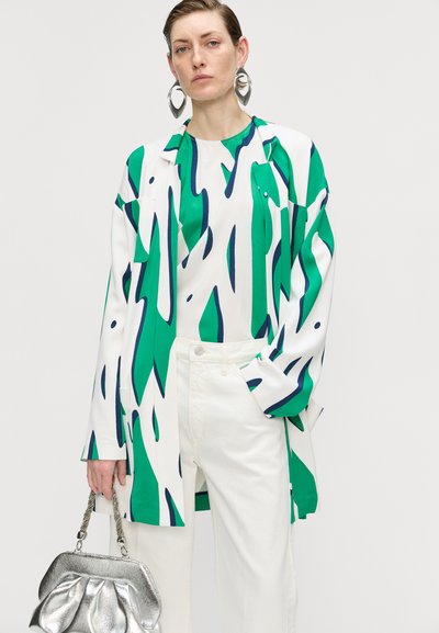Camisa de manga larga con un patrón abstracto en verde y blanco, combinada con pantalones blancos. El modelo sostiene un bolso de mano plateado con un acabado texturizado.