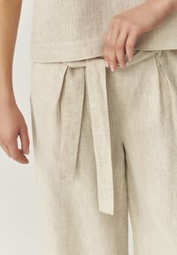 Lichtbeige linnen top en broek met een geknoopte tailleband. De stof heeft een gestructureerde afwerking en de outfit heeft subtiele plooien.