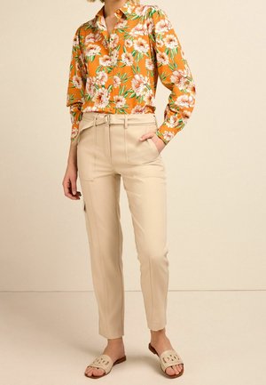 Femme portant une chemise orange à fleurs à manches longues, un pantalon beige taille haute ceinturé, et des sandales beiges à enfiler, debout avec une main dans la poche.