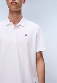 Fehér pamut galléros póló, háromgombos rátéttel és egy kis piros, kék és fehér hímzett logóval a bal mellrészen.