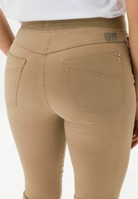Jean taille haute beige avec une ceinture élastique, deux poches arrière et des clous décoratifs sur une poche. Texture de tissu lisse.