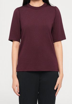 Kobieta ubrana w prostą, krótkim rękawem, ciemno bordową bluzkę z okrągłym dekoltem oraz czarne spodnie, stojąca na jednolitym białym tle.