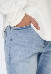 Jeans de color azul claro hechos de mezclilla, con un diseño clásico y costuras visibles. Una mano está colocada en la zona del bolsillo delantero.
