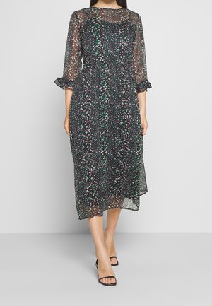 Kobieta w czarnej sukience midi z przezroczystą tkaniną w kwiatowy wzór, rękawy 3/4, mankiety z falbankami oraz czarne buty na wysokim obcasie z odkrytymi palcami na jednolitym tle.