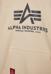 Felpa beige con un grande logo stampato nero, caratterizzato da forme angolari e testo. Include una tasca frontale e un piccolo etichetta rossa sull'orlo.