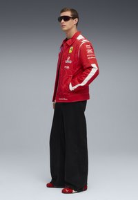 Giovane uomo che indossa una giacca da corsa rossa Ferrari, pantaloni larghi neri, scarpe Puma rosse e grandi occhiali da sole neri, in piedi di lato.