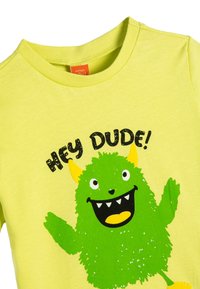 Keltainen puuvillapaita, jossa on vihreä monsterigraafikka ja musta teksti "HEI KAVERI!" sekä keltaiset yksityiskohdat monsterin korvissa ja jaloissa.