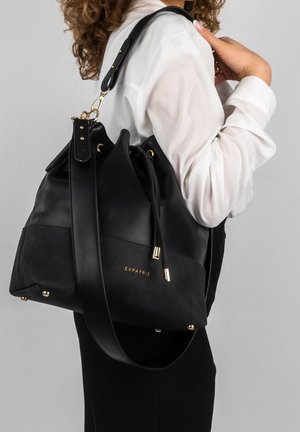 Femme portant une blouse blanche et un pantalon noir, tenant un grand sac à bandoulière en cuir et daim noir avec des accents dorés et le logo "EXPATRIÉ".